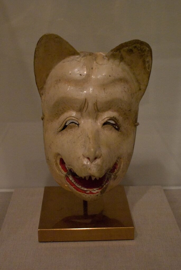 Pale historical fox mask with pointed ears and a red mouth displayed in a museum case